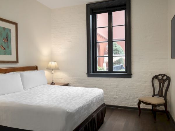 A cozy bedroom with a bed, chair, lamp, window, and wall art on a white brick wall, creating a simple and elegant atmosphere.