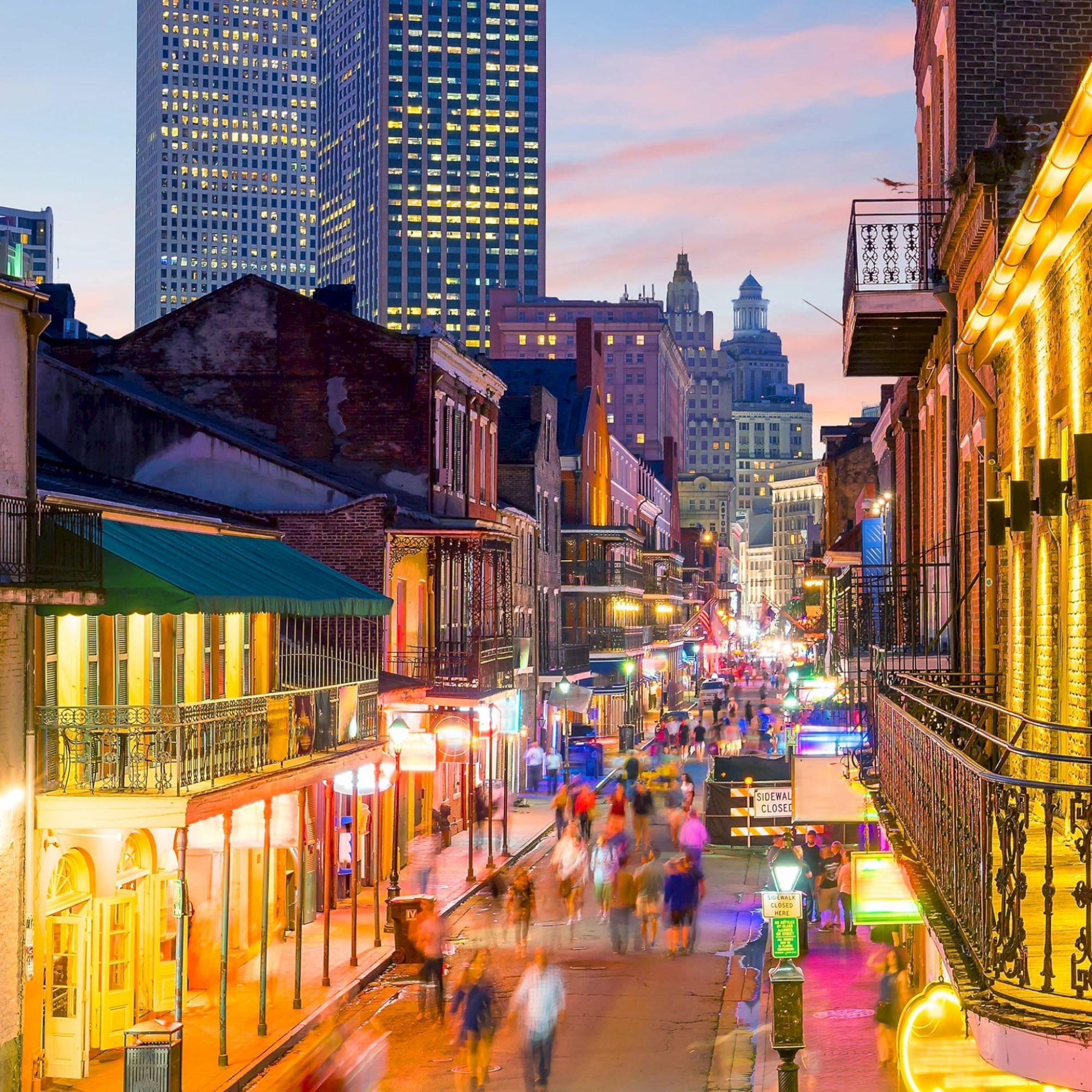 A vibrant, bustling street scene with bright lights, historic buildings, and a crowded pedestrian area under a colorful evening sky.