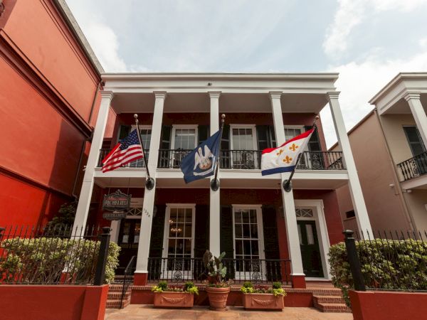 A two-story building with three flags displayed, featuring white columns, red walls, and a gated entrance with potted plants.