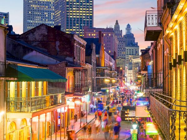 A bustling city street scene with colorful lights and classic architecture, possibly New Orleans, at dusk with people walking around.