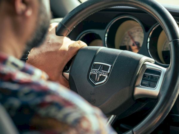 A person is driving a car, holding the steering wheel with a Dodge logo, wearing a patterned shirt.