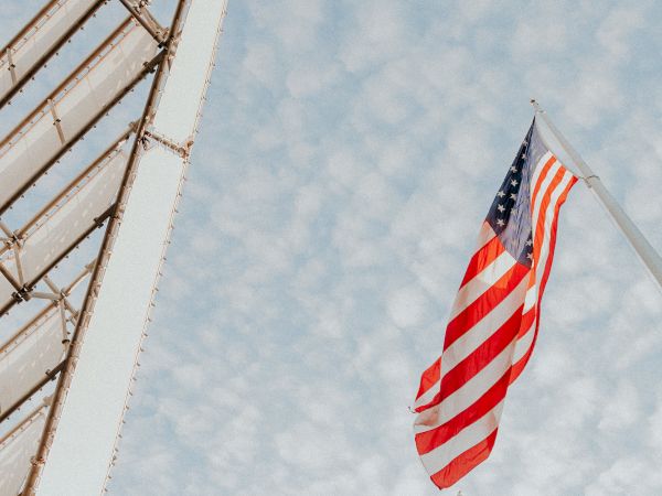 An American flag flies on a pole against a cloudy sky, with a metal structure visible on the left side of the image.