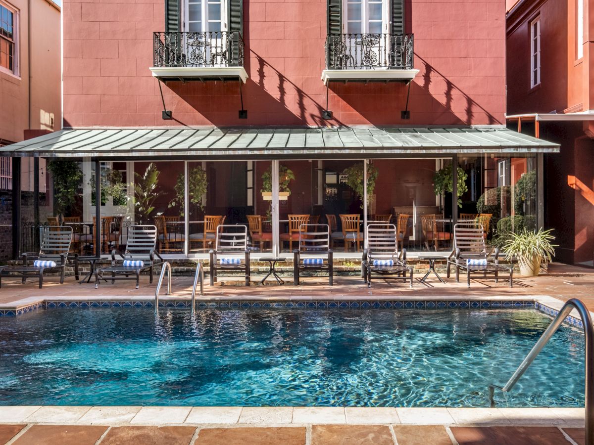 A swimming pool area with lounge chairs, tables, and a building with balconies in the background, creating a relaxing outdoor setting.