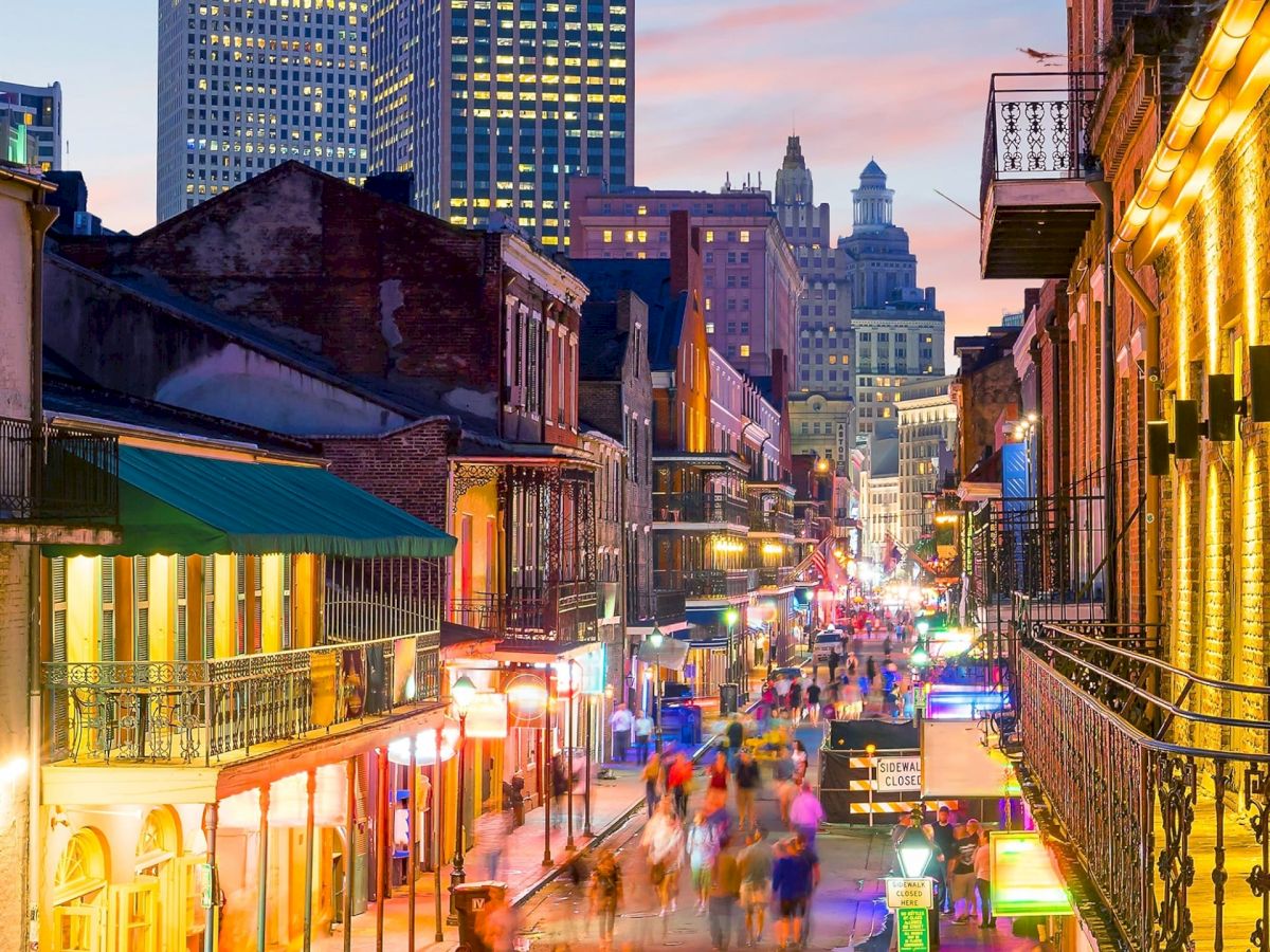 The image shows a lively street scene with colorful lights and blurred crowds, set against a backdrop of tall buildings at twilight.