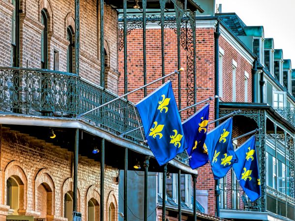 A row of brick buildings with iron balconies displays blue flags featuring yellow fleur-de-lis designs.