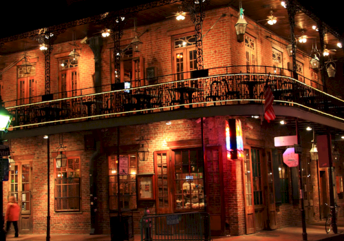 A brick two-story building at night with ornate iron balcony, warm lights, and a corner street scene; people and shopfronts glow softly.