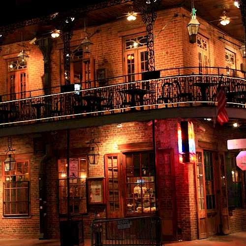 A brick two-story building at night with ornate iron balcony, warm lights, and a corner street scene; people and shopfronts glow softly.