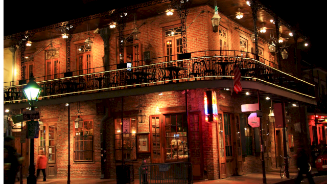A brick two-story building at night with ornate iron balcony, warm lights, and a corner street scene; people and shopfronts glow softly.