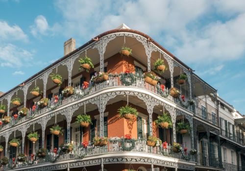A historic building with ornate ironwork balconies and hanging plants, set against a blue sky with clouds.