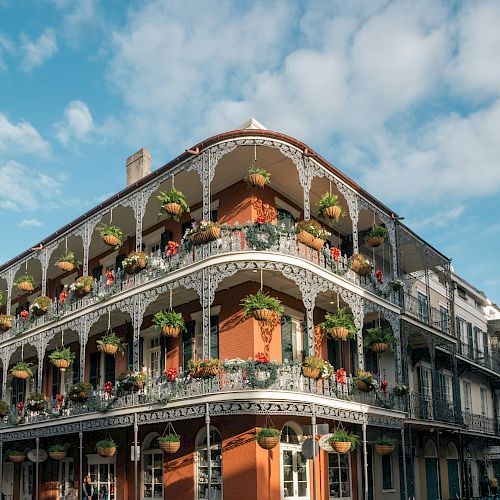 A historic building with ornate ironwork balconies and hanging plants, set against a blue sky with clouds.