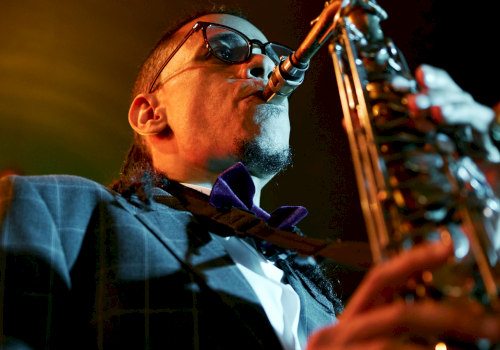 A jazz saxophonist wearing sunglasses and a suit, playing passionately into a microphone on stage under warm lights.