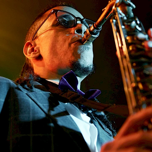 A jazz saxophonist wearing sunglasses and a suit, playing passionately into a microphone on stage under warm lights.