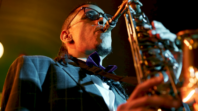 A jazz saxophonist wearing sunglasses and a suit, playing passionately into a microphone on stage under warm lights.