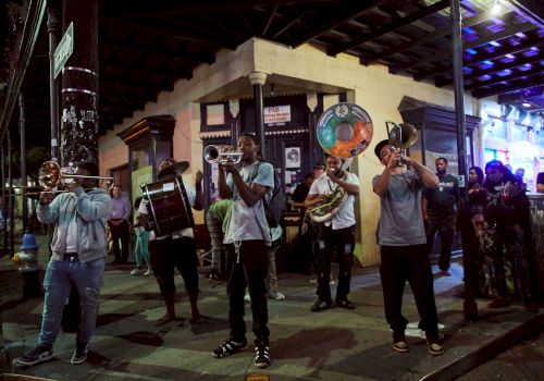 A street marching band plays brass instruments at night under a covered corner, with onlookers and a lit building facade in the background.