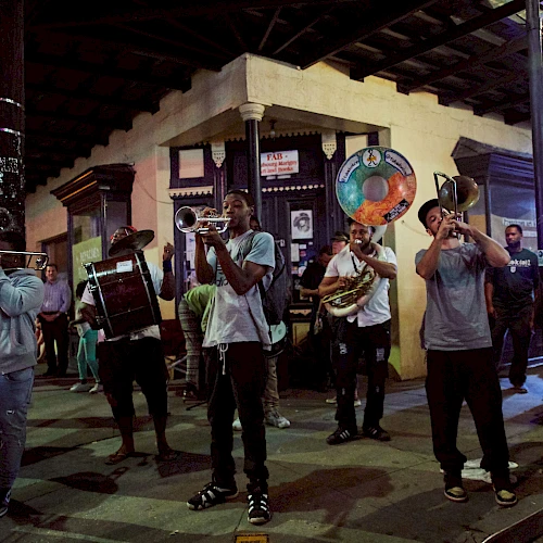 A street marching band plays brass instruments at night under a covered corner, with onlookers and a lit building facade in the background.
