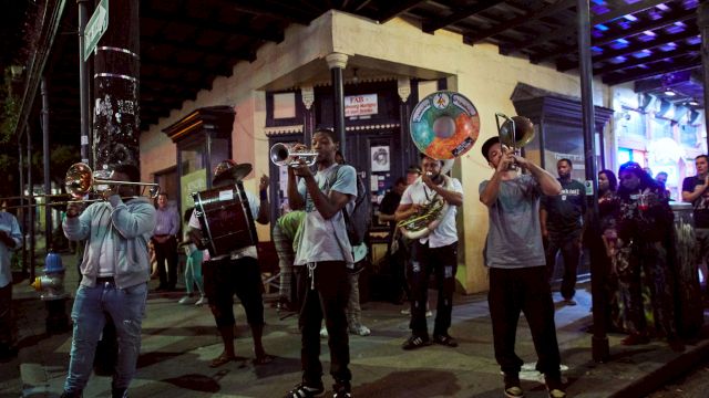 A street marching band plays brass instruments at night under a covered corner, with onlookers and a lit building facade in the background.