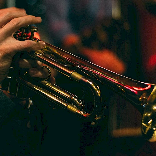 The image shows a person playing a trumpet, with a blurred background suggesting a musical performance or practice setting.