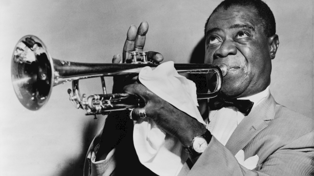 A black-and-white photo of a jazz trumpeter in a suit, playing his horn with a white napkin, smiling while bowing the notes.