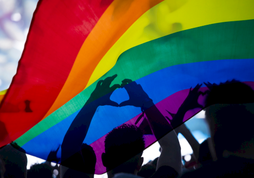 A large rainbow flag is raised in a crowd, silhouettes of people forming heart shapes with their hands under the flag, celebrating Pride.