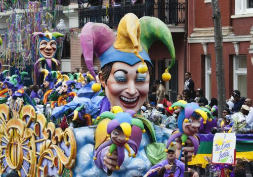 A colorful Mardi Gras parade float features a large jester head, surrounded by people and festive decorations in a vibrant street setting.
