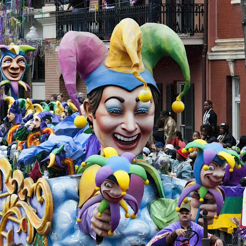 A colorful Mardi Gras parade float features a large jester head, surrounded by people and festive decorations in a vibrant street setting.