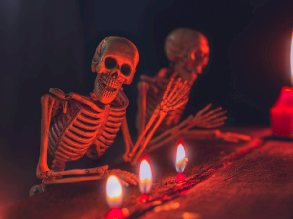 Two skeletons are sitting and leaning on a surface, with candles lit nearby, creating an eerie atmosphere in dim light.