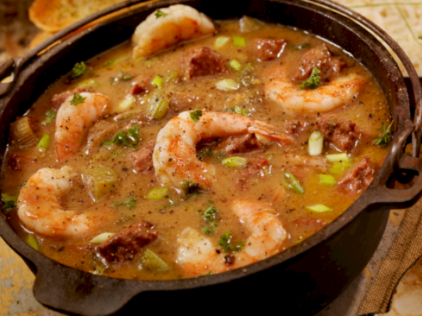 A delicious pot of gumbo with shrimp, sausage, and vegetables in a savory broth is served, garnished with green onions and herbs.