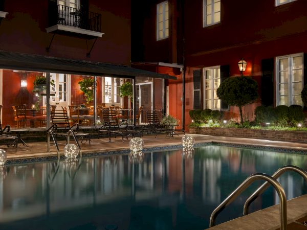 A dimly lit pool area with loungers, illuminated by globe lights, next to a plant-decorated patio with tables and chairs against a red building.