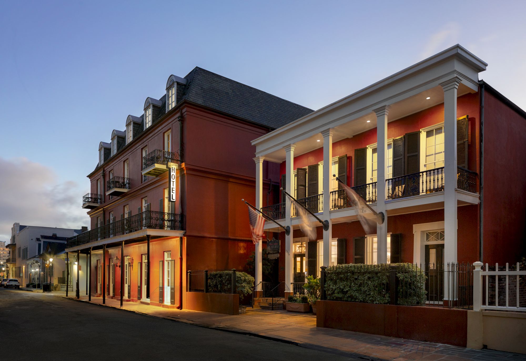 A charming hotel with red walls, balconies, and flags displays a "HOTEL" sign at dusk on a quiet street.