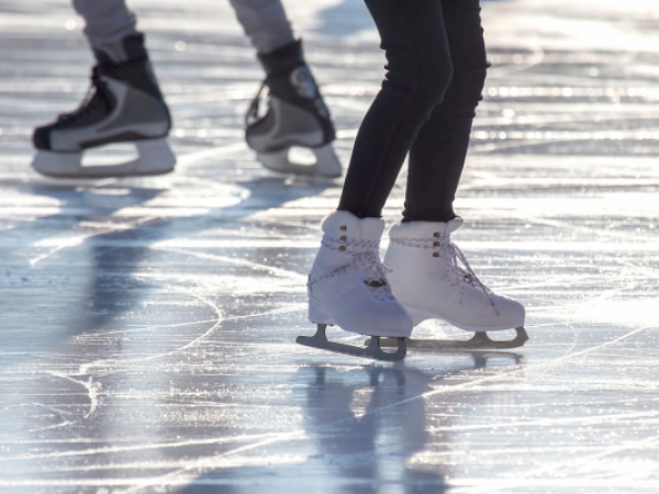 Two people ice skating on a shiny outdoor rink, wearing black and white skates, with reflections visible on the ice.