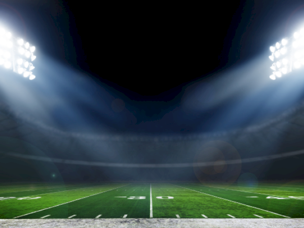 A brightly lit stadium with an empty football field under large floodlights, casting light beams onto the green turf.