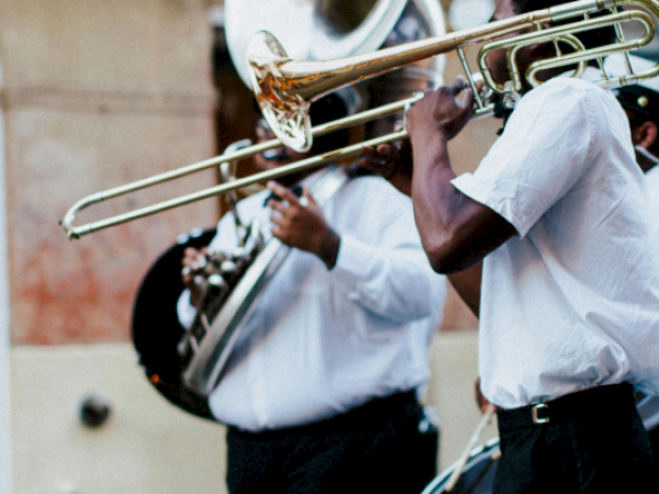 Musicians are playing brass instruments like trombones and tubas, dressed in white shirts, performing outdoors.