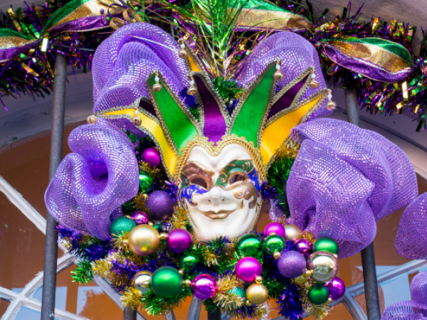 A colorful Mardi Gras mask with green, purple, and gold decorations, including beads and ribbons, is shown hanging.