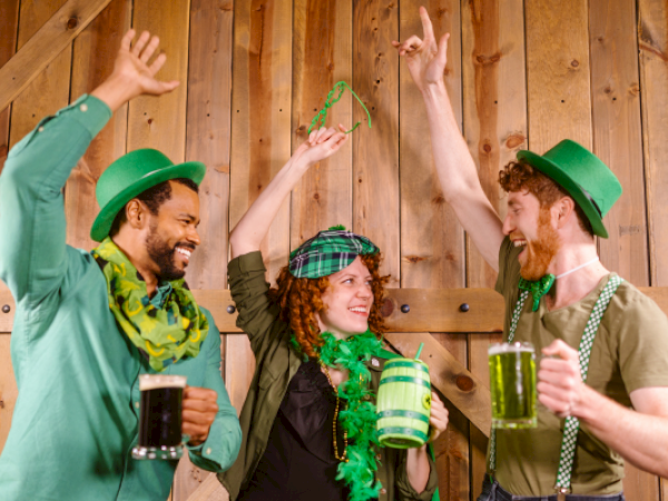 Three people celebrating in green attire, holding mugs with green beer, wearing hats, and smiling joyfully in a festive setting.