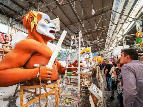 A man wearing an orange and white mask works on a sculpture with a knife, in a busy workshop filled with sculptures and onlookers.
