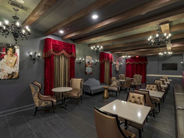 A stylish lounge with dark walls, red drapes, chandeliers, and small tables surrounded by upholstered chairs and booths.