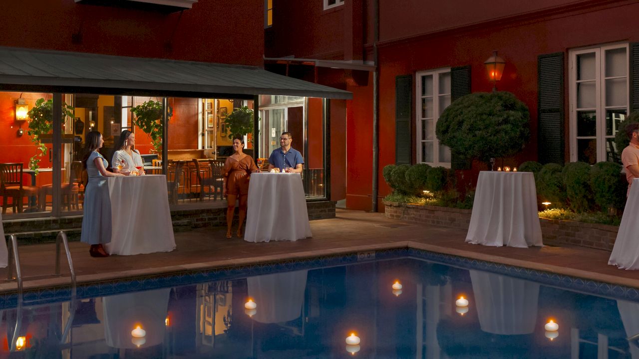 a poolside scene at night with people standing by tables with white tablecloths and candles on them