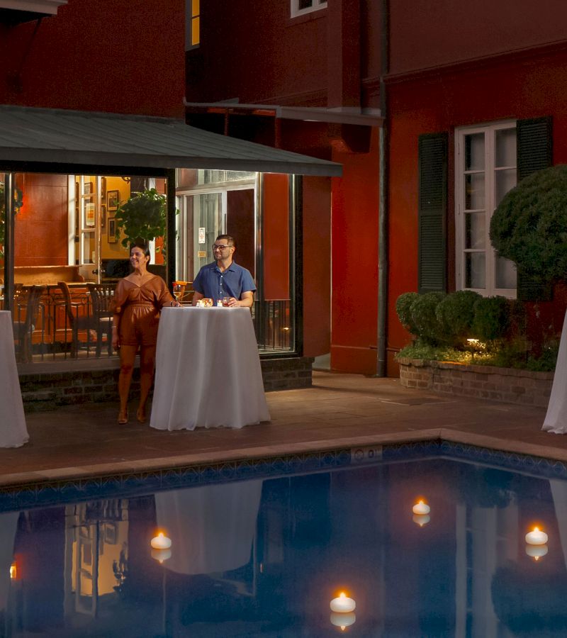 A couple is standing by a pool at night, with a red building in the background.