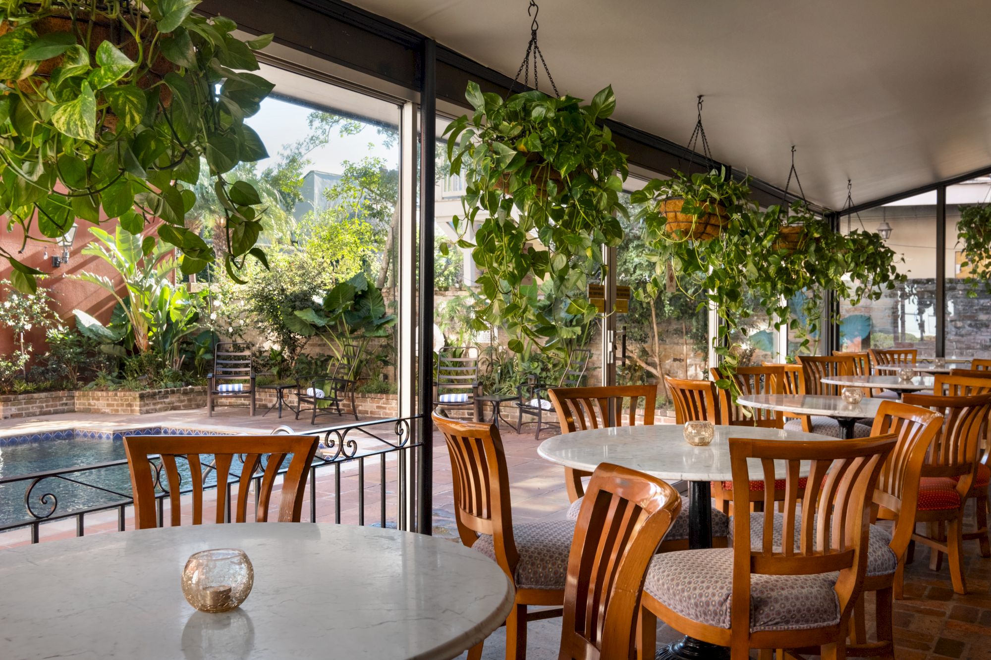 A bright outdoor dining area with round white tables, wooden chairs, hanging green plants, and a railing by a pool or water feature under a covered patio.