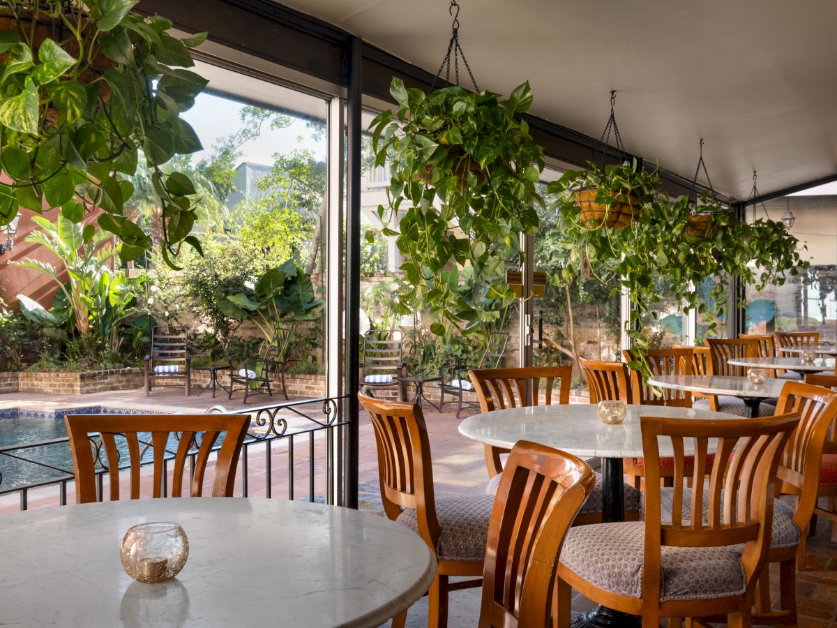 An outdoor dining area with round marble tables, wooden chairs, hanging greenery, potted plants, and a view of a pool or water feature nearby, under a shaded canopy.