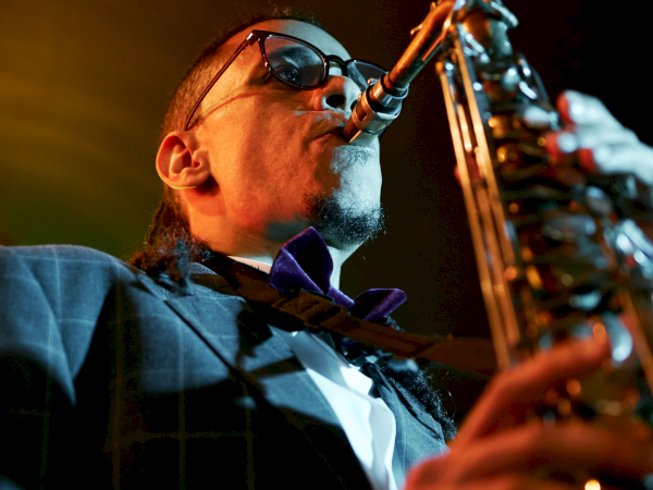 A saxophonist in a suit and sunglasses plays a brass instrument on stage, lit by warm stage lights.