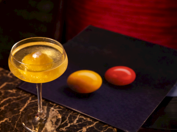 A cocktail glass with a yellow drink sits beside two colored eggs (orange and red) on a dark surface with a vinyl record partially visible, creating a classy still life.