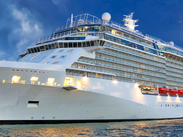 A massive white cruise ship docked at sunset, multiple decks with glass balconies and bright exterior lights illuminating the hull.