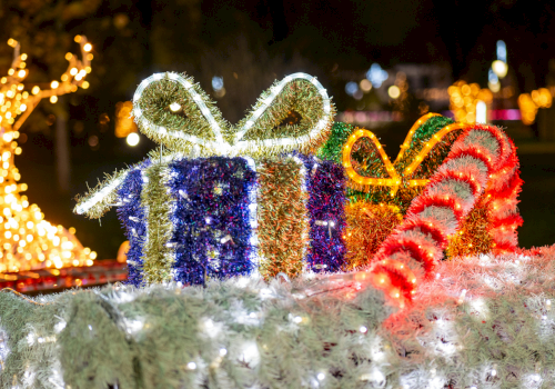 Festive holiday lights form a glowing display with wrapped gifts, candy canes, and a bright decorative bow in an outdoor scene at night.