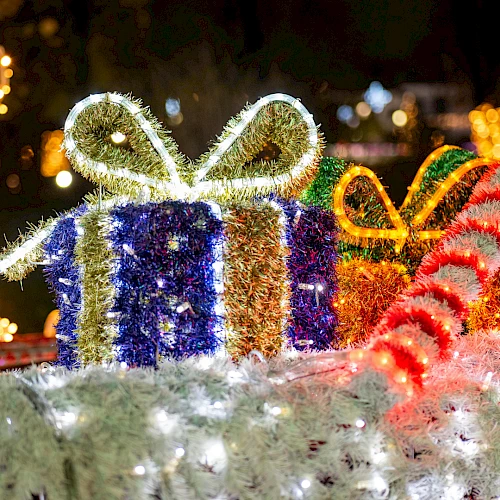 Festive holiday lights form a glowing display with wrapped gifts, candy canes, and a bright decorative bow in an outdoor scene at night.