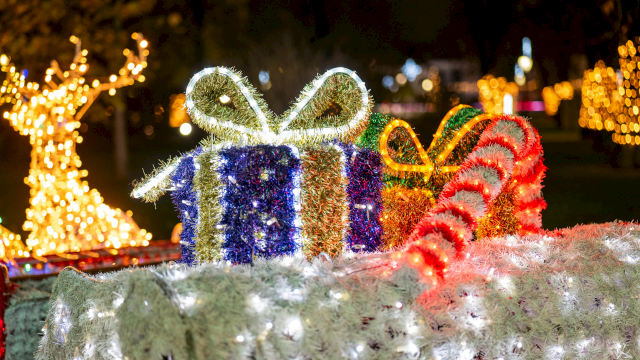 Festive holiday lights form a glowing display with wrapped gifts, candy canes, and a bright decorative bow in an outdoor scene at night.
