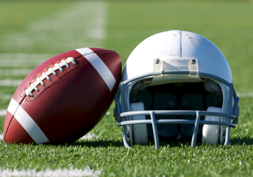 A football and a helmet resting on a grassy field, ready for a play, with white yard lines in the background.