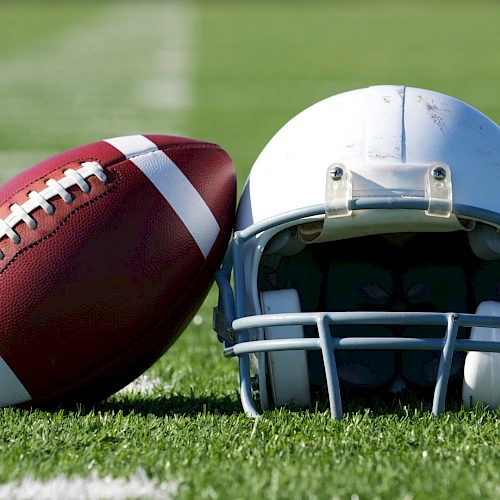 A football and a helmet resting on a grassy field, ready for a play, with white yard lines in the background.
