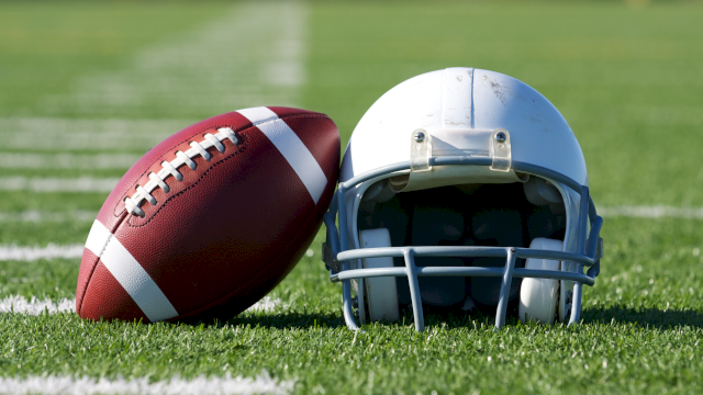 A football and a helmet resting on a grassy field, ready for a play, with white yard lines in the background.