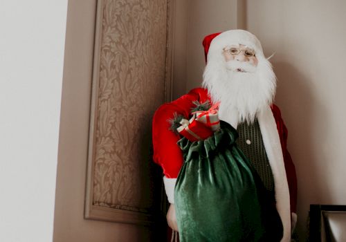 Santa Claus in a red suit carrying a green sack, standing in a doorway with beige walls and patterned paneling.